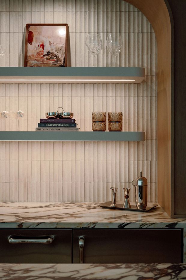 Close-up interior view of a refined built-in bar or drinks cabinet. The composition features vertically fluted light wall panelling, two teal-green floating shelves with integrated linear lighting, a dark timber or painted cabinet base, and a richly veined marble benchtop. Glassware, books, decorative vessels, artwork, and a polished chrome cocktail shaker are arranged on the shelves and counter. A curved timber arch frames the right side, creating a warm, sculptural and luxurious atmosphere.