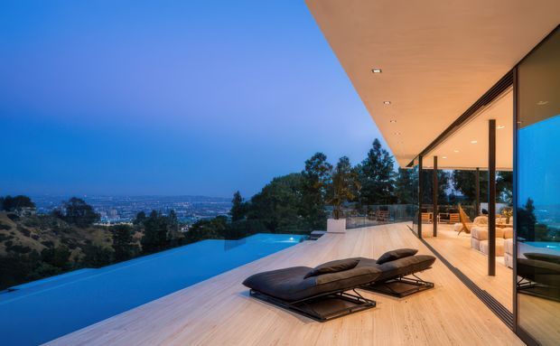 A modern elevated terrace overlooking a broad cityscape, featuring a long infinity-edge pool, pale timber decking, dark sculptural sun loungers, and a glazed living area beneath a deep roof overhang. Warm interior lighting contrasts with the cool blue evening sky, and floor-to-ceiling glazing creates a seamless indoor-outdoor connection.