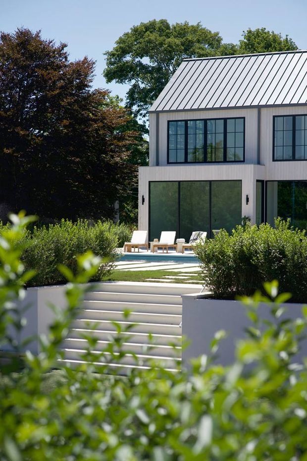 Exterior view of a contemporary two-storey residence set within landscaped gardens. The home has pale vertical cladding, a standing-seam metal gable roof, large black-framed gridded windows, and expansive dark glazed doors opening toward a terrace and swimming pool. Broad white steps and clipped shrubs lead through the garden to the pool area.