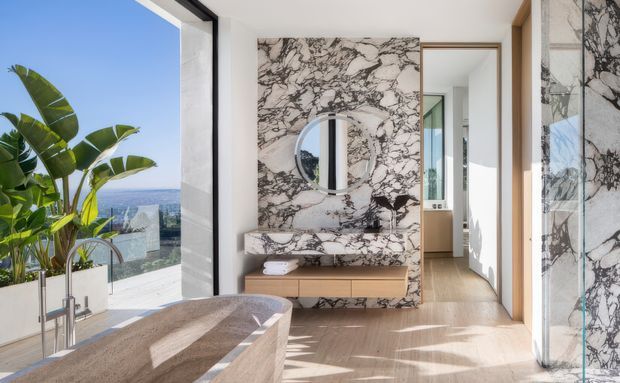 Bright, refined contemporary interior with a freestanding stone bathtub in the foreground, a dramatic veined marble feature wall and matching built-in console, and floor-to-ceiling glazing opening toward a coastal view. Light timber flooring, pale walls, black-framed glazing, and warm timber detailing create an airy luxury bathroom setting, with a hallway or adjacent room visible beyond.
