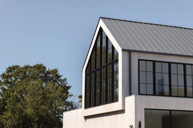 Exterior view of a contemporary two-storey building with a steep gabled roof, light vertical cladding, dark metal-framed grid windows, and a dramatic glazed gable end. A mature tree and clear sky form the background.