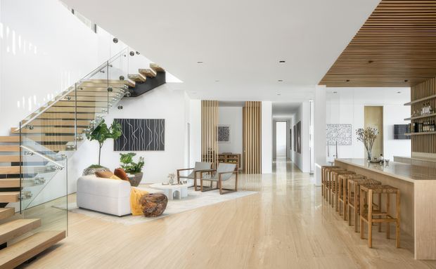 Bright, open-plan contemporary interior featuring a timber staircase with clear glass balustrades, pale timber flooring, a white lounge area, vertical timber screen detailing, and a built-in bar or kitchen counter with timber stools. The space connects a living area, circulation zone, and bar-like entertaining area beneath a slatted timber ceiling feature.