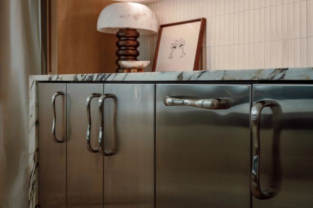 Close-up of a stylish built-in cabinet or sideboard with brushed metallic fronts and sculptural chrome handles, topped by a veined marble slab. A sculptural table lamp and framed artwork sit above it against warm timber panelling and a softly patterned wall.