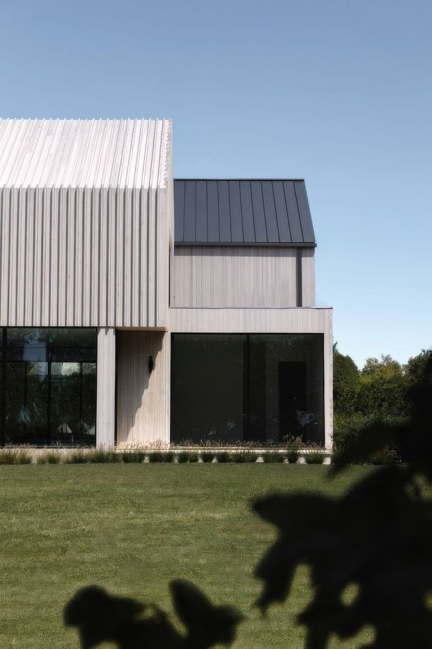 Exterior view of a contemporary, two-level residential building with pale vertically ribbed cladding, dark standing-seam metal roofing, and large dark-framed glazed openings. The building sits behind a manicured lawn and low planting, under a clear blue sky. Strong geometric massing and contrasting roof forms define the facade.