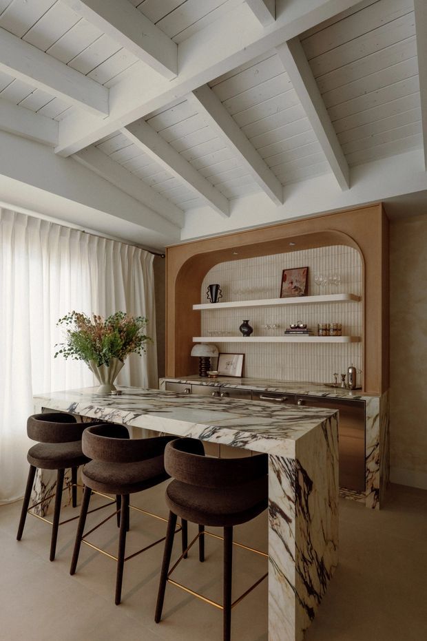 A refined open-plan kitchen and dining area with a large veined marble waterfall island, four upholstered bar stools with brass foot rails, and a built-in timber display and kitchenette wall. The cabinetry features a rounded arch surround, vertically textured light wall lining, open shelves, and integrated drawers. Soft sheer curtains, a sculptural floral arrangement, and a bright white painted exposed-beam ceiling create a warm, elegant atmosphere.