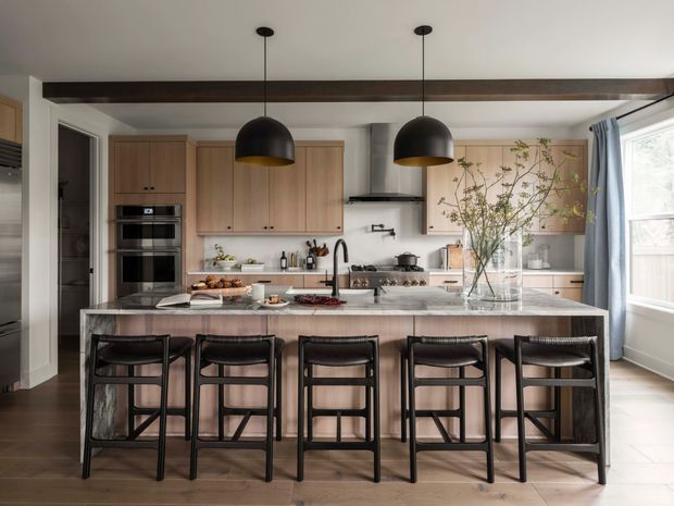 A spacious contemporary kitchen viewed from the dining or living side, featuring a large marble island with five black bar stools, light timber cabinetry, integrated appliances, a gas cooktop, and a stainless-steel rangehood. Two black dome pendant lights hang from a ceiling beam, with a large window and pale blue curtains at right. The room has a warm, refined palette of timber, white, grey, and black.