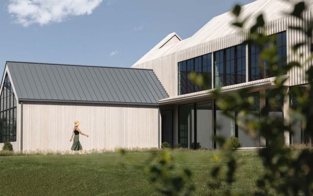Exterior view of a contemporary multi-volume building with pale vertical cladding, dark standing-seam pitched roofs, expansive black-framed glazing, and a covered upper-level terrace or balcony. The structure sits beside a broad lawn, with foreground planting partially framing the image.