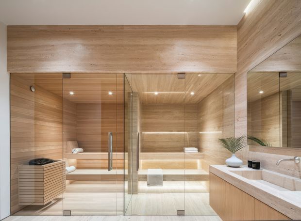 A luxurious contemporary bathroom with extensive warm timber wall and ceiling lining, a glass-enclosed sauna or steam room on the left, a freestanding bathtub in a lit alcove at the rear, and a timber vanity with a pale stone benchtop and integrated basin on the right. Large mirrors, frameless glass panels, concealed lighting, and clean linear detailing create a refined spa-like interior.