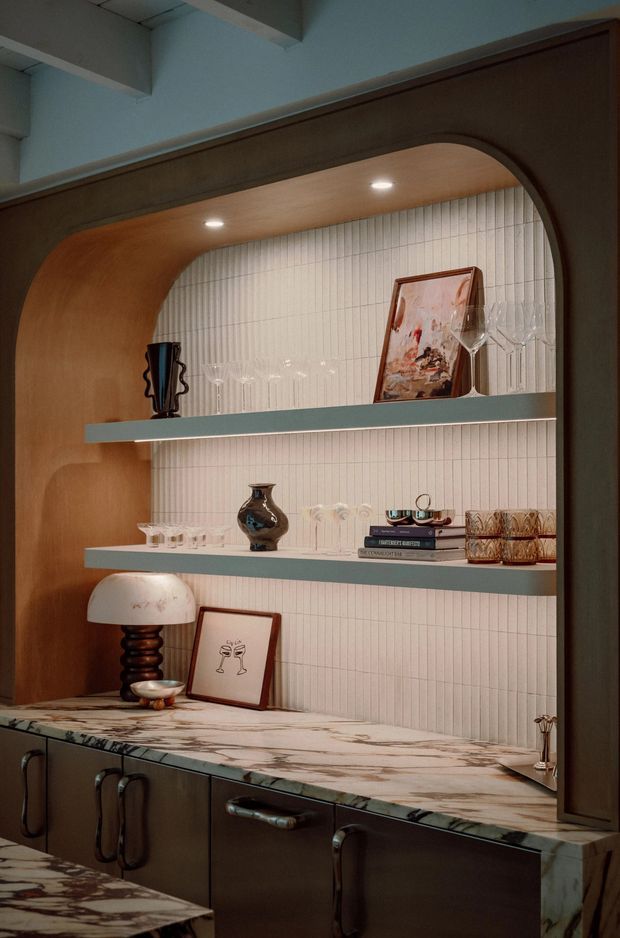 A close-up interior view of a sophisticated built-in drinks bar or display cabinet. A dark timber arched surround frames a vertically ribbed white tile backsplash, with two pale shelves displaying glassware, books, artwork and decorative objects. The lower section has dark cabinetry with sculptural metal pulls and a richly veined marble benchtop. Warm recessed lighting and curved detailing create a refined, intimate atmosphere.
