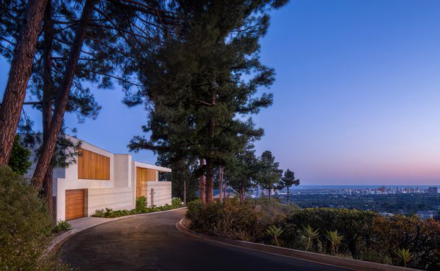 Twilight exterior view of a modern multi-level residence set among tall pine trees on a sloping site. The home has clean geometric white rendered walls, warm timber-clad garage and window panels, and broad glazing. A curved driveway and landscaped foreground lead toward an expansive city and coastal horizon view.