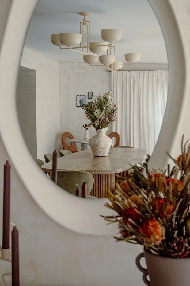 A warm, softly styled dining room viewed through a large rounded organic-shaped wall mirror. The reflected room features a round marble or stone dining table with a fluted timber base, curved upholstered dining chairs, a sculptural cream vase with dried flowers, sheer curtains, textured pale walls, framed artwork, and a contemporary multi-arm pendant light. Dried floral arrangements and taper candles sit in the foreground, adding an earthy decorative layer.