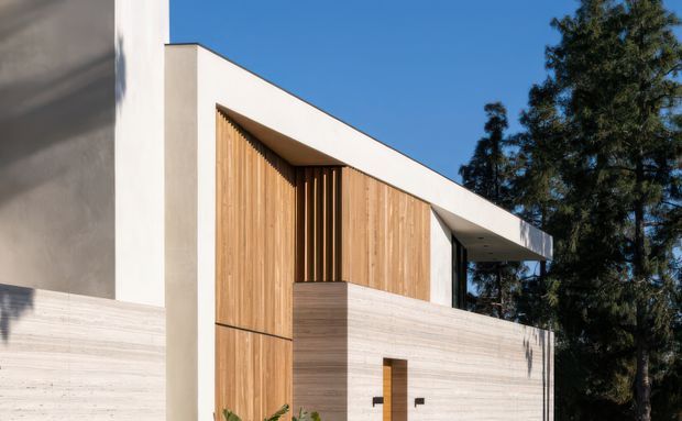 Modern residential facade viewed from outside, featuring a crisp white rendered upper structure and projecting flat roof canopy, warm vertical timber cladding and screening, and a pale horizontal-textured masonry or concrete boundary wall. A minimalist timber entry door is visible, with mature trees behind the building.