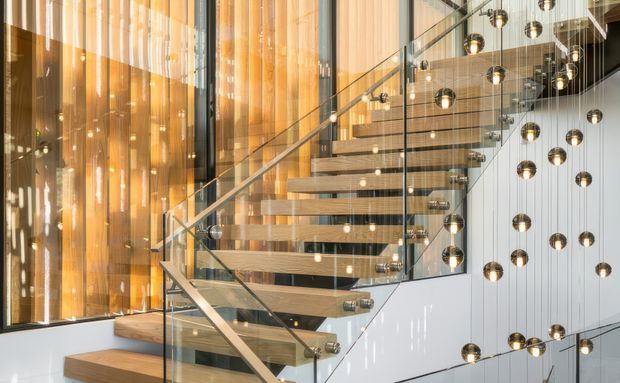 Contemporary interior stairway featuring floating timber treads, clear glass balustrades, slim metal handrails, and a dramatic installation of suspended spherical pendant lights. Warm timber wall cladding and abundant glazing create a refined, light-filled architectural space.