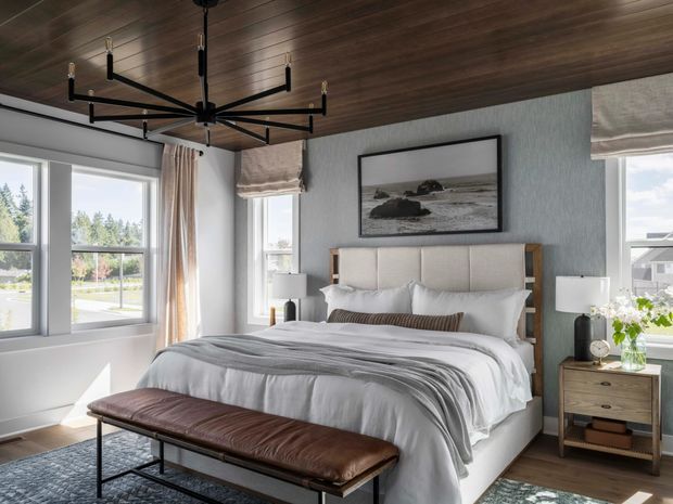 A bright, refined bedroom with a large upholstered bed, layered white and grey bedding, bedside tables with lamps, and a long brown leather bench at the foot of the bed. The room features a dark timber-plank ceiling, pale textured walls, large windows with Roman blinds and curtains, and a black linear chandelier. A monochrome coastal landscape artwork hangs above the bed.