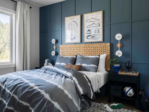A stylish bedroom with a neatly made bed against a dark blue paneled feature wall. The room includes a woven timber headboard, blue and white bedding, black bedside furniture, graphic framed artwork, globe-like wall sconces, white curtains, and sports accessories. Natural light enters through the large window on the left.