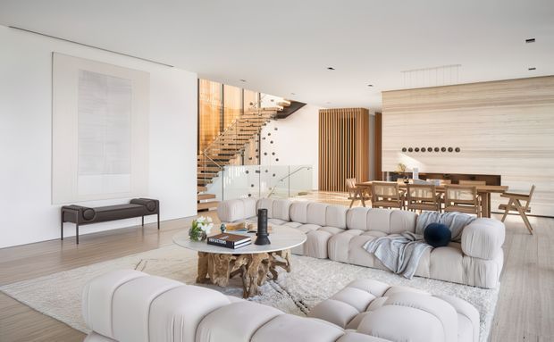 A bright, spacious open-plan living and dining area with a contemporary minimalist interior. The room features sculptural cream modular sofas, a round coffee table, a neutral textured rug, light timber flooring, a large abstract wall artwork, a glass balustraded staircase, vertical timber screens, and an integrated dining and kitchen area with extensive light timber cabinetry. Recessed ceiling lighting and abundant neutral finishes create an airy, refined atmosphere.