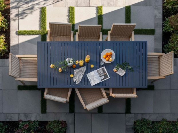 Overhead view of a contemporary outdoor dining setting arranged on a paved terrace. A long dark slatted dining table is surrounded by woven outdoor chairs, with place settings, fruit, flowers and reading materials on the tabletop. Large pale rectangular paving slabs are bordered by clipped green planting and hedges, creating a formal landscaped courtyard atmosphere.