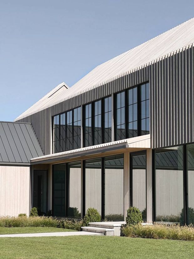 Exterior architectural photograph of a contemporary building with a steeply pitched light metal roof, vertically ribbed metal cladding, extensive black-framed floor-to-ceiling glazing, and a glazed entrance canopy. The building sits within a neatly landscaped lawn with low ornamental planting under a clear blue sky.