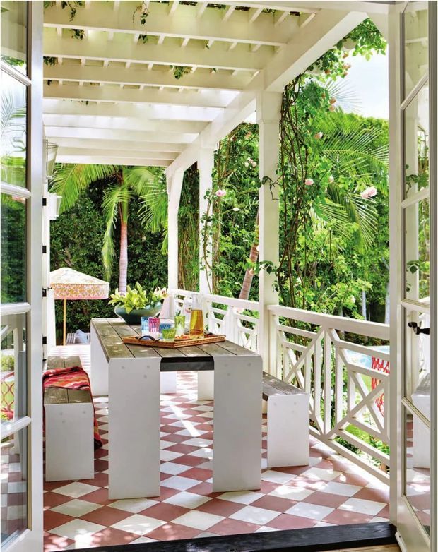 Bright tropical covered outdoor dining veranda viewed through open French doors. The space features a white-painted timber pergola with exposed rafters, white posts and cross-braced balustrades, lush climbing and tropical vegetation, a rustic timber dining tabletop on white block-style legs, and a red-and-white geometric checkerboard tiled floor. The open, airy setting is strongly connected to the garden.