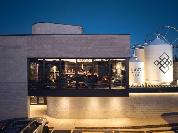 Twilight exterior view of a contemporary whisky house or distillery, with a rectilinear pale brick/concrete-block facade, dark-framed floor-to-ceiling glazing revealing a warmly lit interior, and large cylindrical whisky tanks prominently displayed on the right. A vehicle and street edge appear in the foreground.