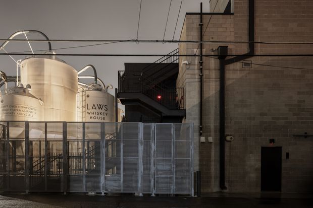 Exterior view of an urban industrial whiskey distillery, showing large stainless-steel fermentation or storage tanks marked “Laws Whiskey House,” a secured metal louvred fence and gate, exposed overhead utility lines, dark metal platforms and stairs, and a substantial brown brick industrial building.