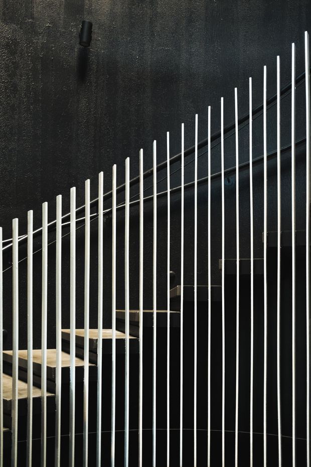 Close-up architectural photograph of a contemporary stairway with pale timber treads and dark stringers, screened by a dense vertical metal balustrade. The surrounding walls and ceiling are dark, textured, and nearly black, creating a strong graphic contrast with the light rails and stair edges. Minimal black lighting and concealed structural details reinforce the dramatic, refined interior aesthetic.