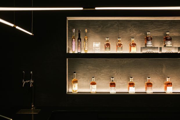 Moody, dark-toned bar interior featuring two long, illuminated recessed display shelves lined with liquor bottles. The shelving is clad in dark timber or wood-grain panels with black surrounding walls and integrated warm linear lighting. A small bar tap and counter are visible in the foreground, while slim geometric pendant or track lights hang overhead.