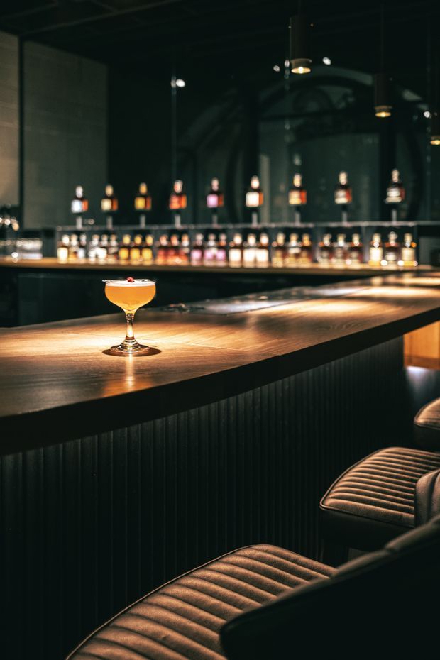 A dimly lit, sophisticated cocktail bar interior with a long warm timber bar counter, dark vertically ribbed bar front, mirrored back bar lined with illuminated liquor bottles, pendant lights, and ribbed upholstered bar stools. A coupe glass cocktail provides a small focal accent on the counter. The atmosphere is intimate, moody, and refined.