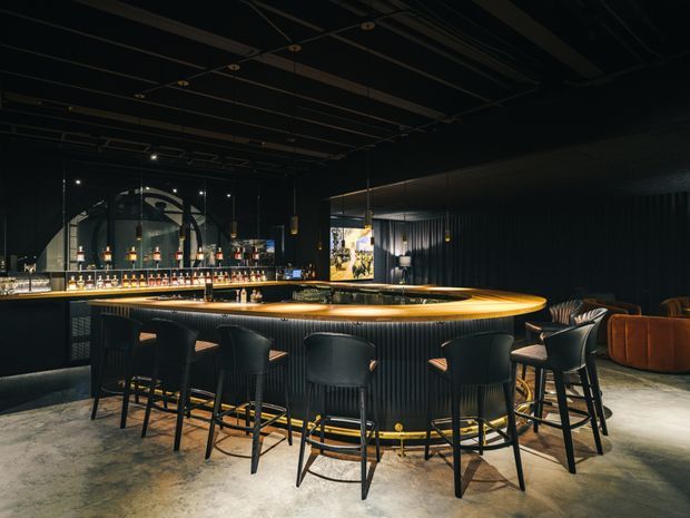 A dark, atmospheric bar interior dominated by a large circular island bar with a warm timber-toned countertop and integrated lighting. Black fluted wall cladding, black ceiling structure, pendant lights, bar shelving, and upholstered bar stools create a dramatic hospitality setting. A polished concrete floor and lounge seating are visible in the background.