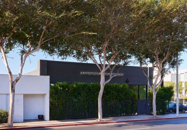 Street-facing view of a low-rise contemporary commercial building, largely obscured by mature trees and neatly trimmed hedges. The facade combines a dark charcoal rectangular volume with a white rendered entrance structure, dark-framed glazing, a metal security fence, and a paved sidewalk with a red-painted curb. A mounted business sign is visible on the dark facade.