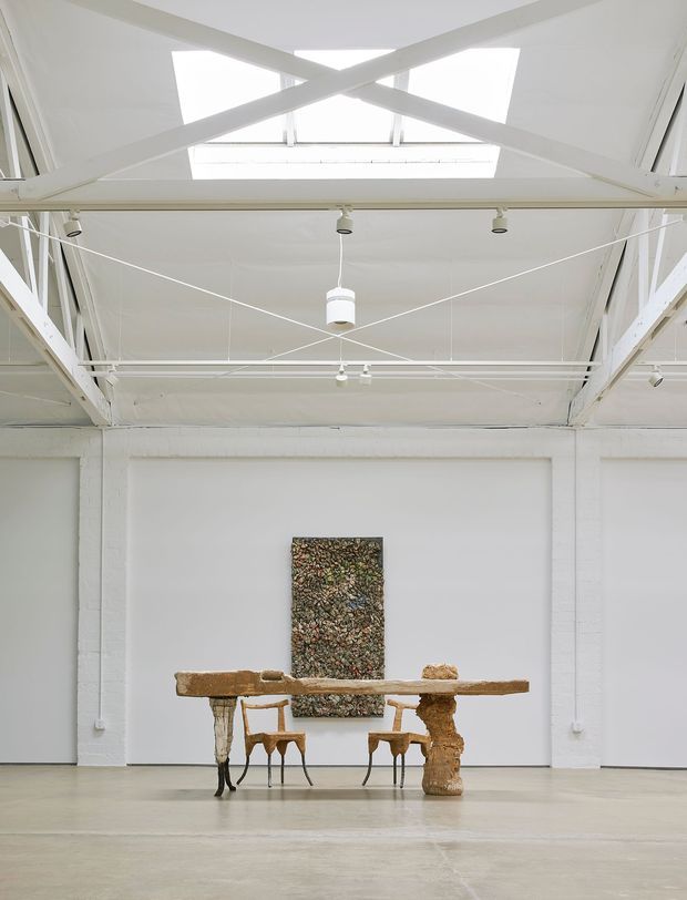 A spacious white-painted gallery or studio interior with exposed structural beams, a high industrial-style ceiling, large skylights, suspended track and pendant lighting, and polished concrete flooring. A rustic sculptural timber table with sculptural supports and two wooden chairs is arranged centrally against a white wall, with an abstract textured artwork behind it.