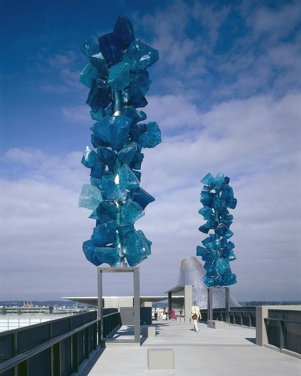 A rooftop pedestrian terrace or public architectural walkway with dark metal balustrades and large blue crystalline sculptural installations mounted on tall rectangular frames. A modern structure and distant urban waterfront skyline are visible beneath a cloudy blue sky.