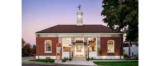 Exterior view of a symmetrical single-storey red-brick civic or commercial building with a low hipped roof, prominent white classical trim, tall arched windows, a central entrance, and a lantern-like cupola. The building fronts a paved sidewalk and is framed by mature trees.