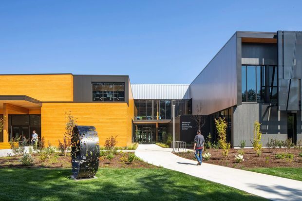 Exterior view of a contemporary multi-building institutional or commercial complex surrounding a landscaped courtyard. The composition includes a warm timber-clad low wing, dark grey and silver metal-clad volumes, extensive floor-to-ceiling glazing, a covered central entrance, paved pedestrian paths, planted garden beds, lawn, and outdoor sculpture. Text on the central facade appears to identify an educational or community centre.