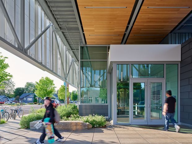 Contemporary glazed building entrance viewed from an exterior pedestrian forecourt. The facade combines extensive floor-to-ceiling glass curtain walling, dark horizontal cladding, light rendered panels, and a warm timber soffit with recessed linear lighting. A glass entry door and vestibule are centered in the frame, with landscaping, bicycles, pedestrians, and neighbouring buildings visible around the approach.