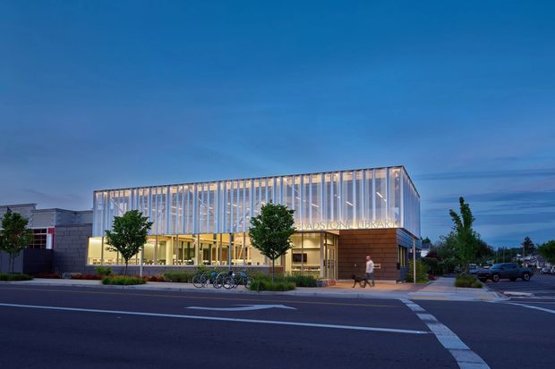 Twilight exterior photograph of a contemporary public library building on an urban street corner. The building has a large transparent glazed upper volume with closely spaced vertical fins, a warm illuminated ground-floor entrance, brick-toned cladding, landscaped edges, bicycles, and marked roads in the foreground.