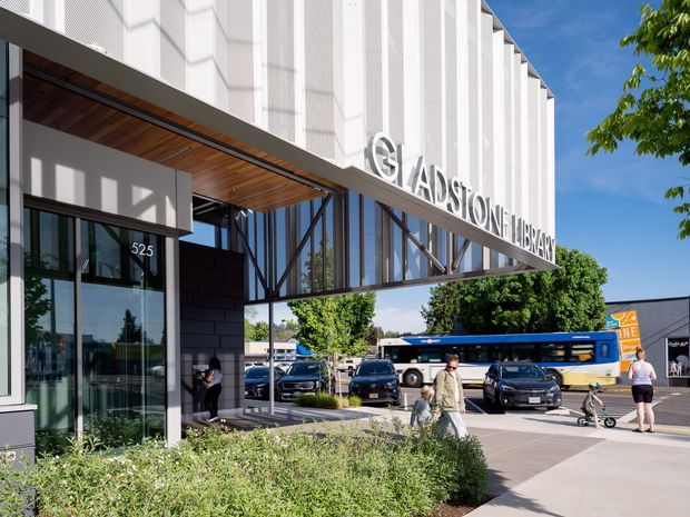 Exterior street-level view of the Gladstone Library, featuring a contemporary civic building with a projecting canopy, tall vertical white facade fins, extensive glazed walls, timber soffit lining, and prominent library signage. The forecourt includes landscaping, pedestrians, parked vehicles, and a public bus.