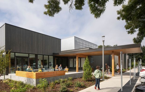 Contemporary single-storey building exterior with a dark vertically clad rectangular volume, a light corrugated-metal upper volume, extensive floor-to-ceiling glazing, and a timber-framed covered entry canopy. The landscaped frontage includes low planting, timber-edged seating or planter elements, pedestrians, and a paved access path. The image appears to show a public or commercial facility rather than a private residence.