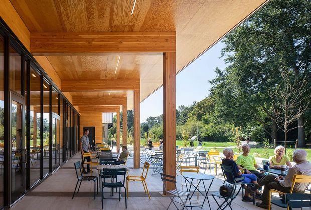 A covered outdoor terrace beside a glazed building facade, featuring a strong rhythm of timber columns and a warm timber-lined soffit. Numerous lightweight tables and chairs are arranged for casual dining or gathering, with people seated at the edge overlooking landscaped grounds. The space is bright, open, and connected to the surrounding garden through extensive black-framed glazing.