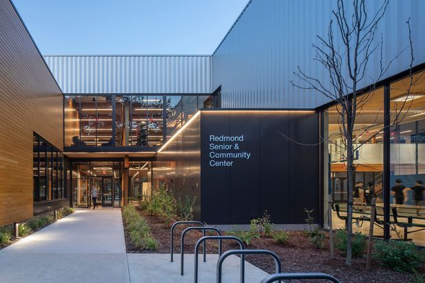Exterior view of a contemporary community centre arranged around a landscaped courtyard. The building features corrugated metal cladding, warm timber-toned soffits and walls, extensive glazed facades, a dark entrance volume with illuminated signage, and a pedestrian path with bicycle racks in the foreground.