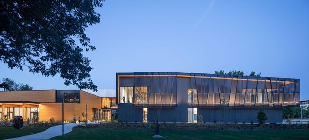 Exterior view of a contemporary low-rise building at dusk, composed of a warm-toned single-storey wing connected to a larger dark-clad rectangular volume. The main building features extensive floor-to-ceiling glazing, a projecting upper-level-like glazed facade, strong horizontal rooflines, and warm interior lighting. Landscaped lawn, mature trees, and a paved path occupy the foreground.