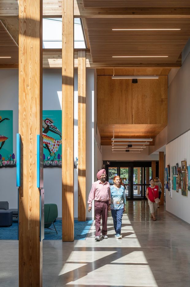 A bright, contemporary institutional corridor or gallery-like interior with a polished concrete floor, extensive exposed timber columns and timber-lined ceilings, linear recessed lighting, clerestory glazing, and framed artwork along the walls. A colorful mural and lounge seating are visible to the left, while several people walk through the spacious hallway. Strong sunlight creates geometric shadows across the floor.
