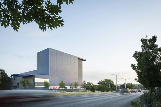 Exterior architectural view of a large contemporary institutional or commercial building beside a broad urban roadway. The building has a strongly geometric form, a tall rectangular volume clad in pale grey metal panels, a deep projecting roof edge, glazed lower-level frontage, and a lower adjoining wing. Street trees and landscaped planting frame the scene, with traffic visible on the road.