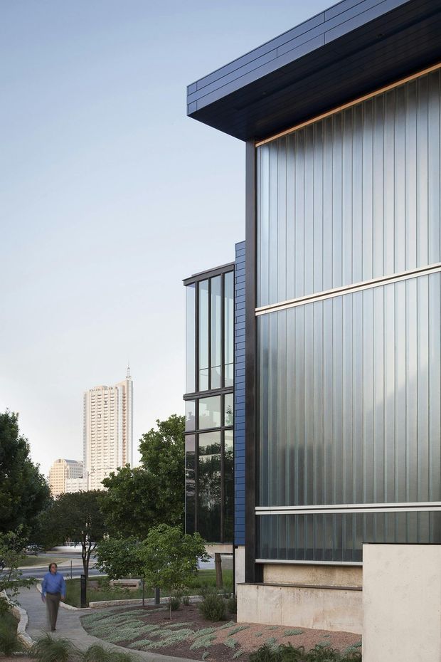 Exterior architectural photograph of a contemporary multi-storey building in an urban, landscaped setting. The facade combines dark metal cladding and trim, vertical translucent ribbed glazing or panels, and tall black-framed windows beneath a deep projecting flat roof. Trees, pedestrian paths, and distant high-rise buildings are visible around the building.