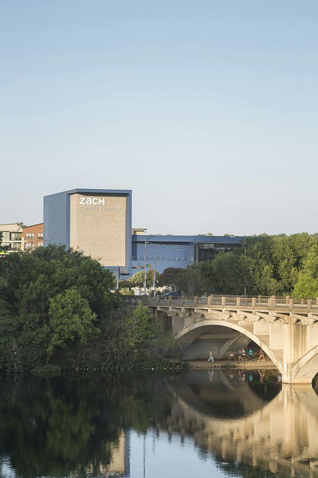 Exterior architectural photograph of the Zach Theatre complex viewed across a calm river, with a prominent rectangular theatre tower bearing the theatre signage, a low modern building extending behind it, mature trees, and a large arched masonry bridge in the foreground. The image has a calm, civic urban setting and soft natural daylight.