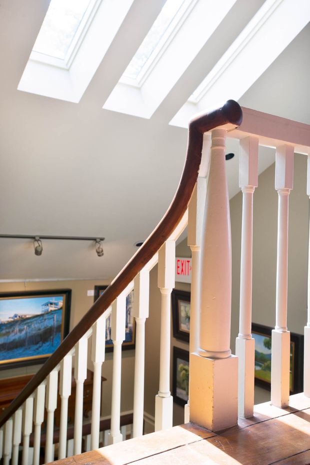 Interior view of a traditional stairway with white painted turned balusters and newel post, a dark stained timber handrail, and warm timber stair treads. Three roof skylights bring daylight into the stairwell, with framed artwork and track lighting visible on the surrounding walls.