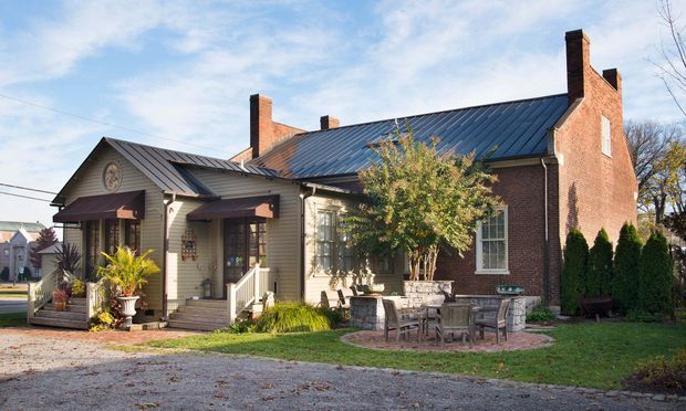 Exterior view of a traditional brick and weatherboard house with a dark metal gabled roof, prominent brick chimneys, a small covered entry porch, timber-framed windows, and a landscaped patio with outdoor dining furniture. The home sits in a suburban or village-like setting.