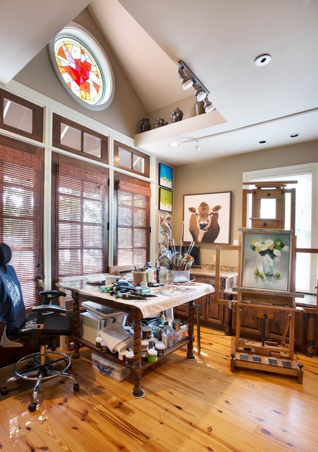 A bright, high-ceilinged artist's studio with warm timber flooring, extensive timber-framed windows fitted with woven blinds, and a distinctive circular stained-glass window. A central worktable is covered with painting supplies, with storage beneath, while framed artwork, shelving, cabinetry, and two easels occupy the room. Track lighting and recessed lights illuminate the creative workspace.