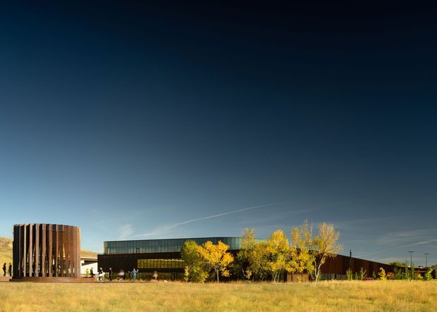 Wide exterior view of a contemporary low-rise building set in an open grassy landscape. The composition features a dark horizontal building volume with extensive glazing, a distinctive tall rounded tower or cylindrical pavilion clad in vertical timber slats, and yellow autumn trees along the frontage. The clear sky and expansive foreground emphasize the building's isolated, low-density setting.