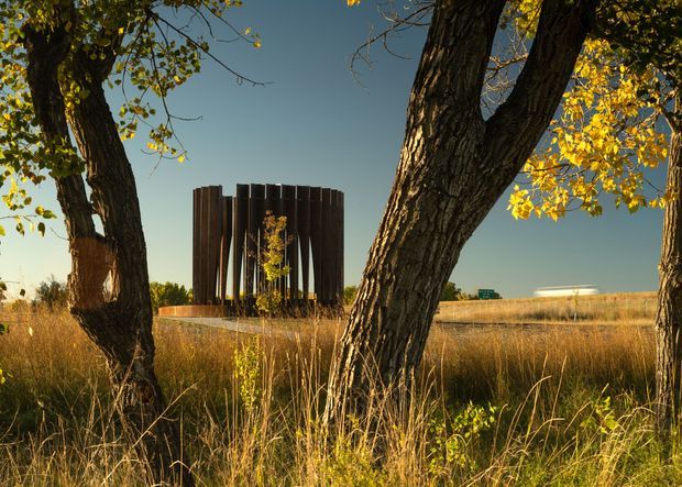 A cylindrical dark-clad architectural structure stands in an open rural landscape, viewed through mature trees and tall golden grasses. The building has a sculptural, vertically ribbed facade and appears to be a contemporary pavilion or small building, with warm low-angle sunlight and autumn foliage creating a dramatic natural setting.